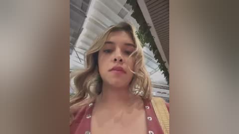 Media: Video of a young blonde woman with wavy hair, wearing a red top, standing in front of a modern building with glass windows and greenery.