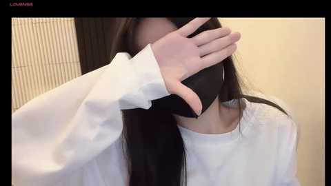 Media: Video of a person with long black hair, wearing a white shirt, and black mask covering their mouth, standing against a beige wall with a light wooden backdrop.