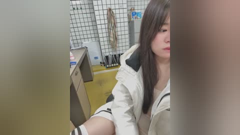 Media: Video of a young Asian woman with long brown hair, wearing a white jacket, sitting at a desk in a small, tiled room with a yellow floor and a shelf of cleaning supplies.