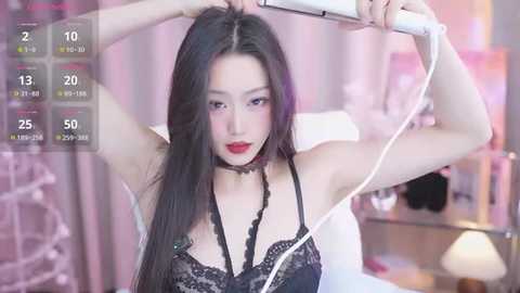 Video of a young East Asian woman with long black hair, red lipstick, and dark lingerie, using a hairdryer in a softly lit bedroom with a white bed and pink curtains.