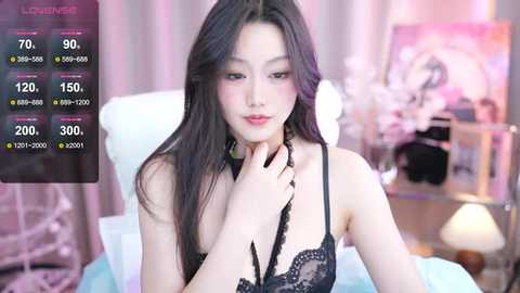 Media: Video of an Asian woman with long black hair, wearing black lace lingerie, seated on a bed with pastel-colored bedding. The background features soft lighting and a blurred, pink-hued room.