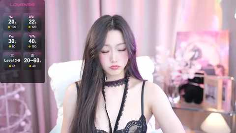 Media: Video of an East Asian woman with long black hair, wearing black lace lingerie, sitting on a bed with white pillows. Background features a soft-focus bedroom with floral decor.