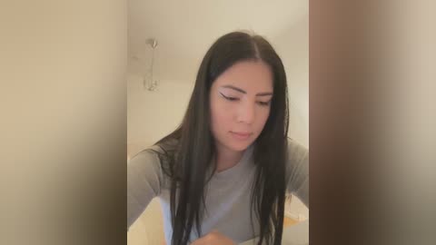 Media: Video of a young woman with long, straight black hair, wearing a gray shirt, sitting in a bathroom with beige walls and a towel rack.