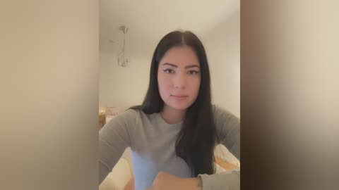 Media: Video of a young Asian woman with long black hair, wearing a gray top, standing in a dimly lit, minimalist bedroom with cream walls and a white bed.