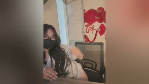 Media: A blurry video of an Asian woman with long black hair, wearing glasses and a mask, leaning over a table in a dimly lit room with floral wallpaper and a red \"F\" sign.