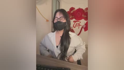 Media: A video of an East Asian woman with long black hair, glasses, and a black face mask, wearing a light gray hoodie, seated at a desk in a dimly lit room with a red floral wall hanging.