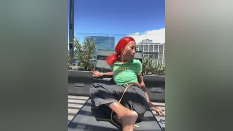 Video of a woman with red hair, wearing a green T-shirt and gray shorts, sitting on a rooftop deck with a cityscape in the background.