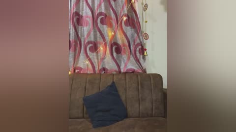 Media: Video of a brown couch with a dark blue pillow, set against a wall adorned with heart-shaped curtains featuring pink and white patterns, and a dreamcatcher hanging on the right.