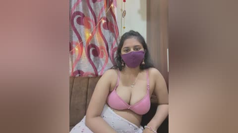 Media: Video of a South Asian woman with medium skin tone and long black hair, wearing a purple mask, pink bra, and floral-patterned skirt, sitting on a couch with a patterned curtain and string lights in the background.