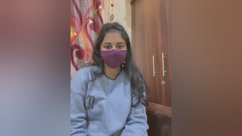 Media: Video of a South Asian woman with long black hair, wearing a purple face mask and light blue sweater, sitting indoors near a brown couch and patterned curtain.