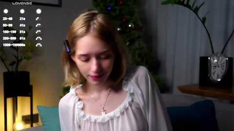 Media: Video of a young Caucasian woman with shoulder-length blonde hair, wearing a white blouse, seated indoors with a dimly lit Christmas tree in the background.