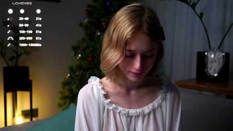 Media: Video of a young blonde woman in a white blouse, standing indoors next to a lit Christmas tree and potted plants.