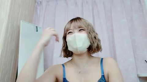 Video of a young Asian woman with short, wavy blonde hair, wearing a blue lace bra and a white face mask, standing in a bathroom with light-colored walls and a pink shower curtain.