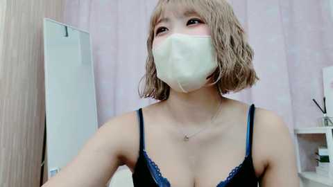 Video of an Asian woman with short, wavy, light brown hair, wearing a black lace bra, face mask, and necklace, standing in a pastel-colored room with a mirror and a white shelf in the background.