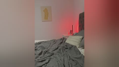 Media: Video of a minimalist bedroom with a neatly made bed covered in gray blankets, a headboard, a yellow abstract painting, and red accent lighting.