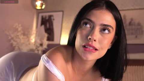 Media: Video of a young woman with long black hair, light skin, and a light pink off-shoulder top, looking contemplative in a warmly lit, softly blurred room with framed photos and a decorative plant.