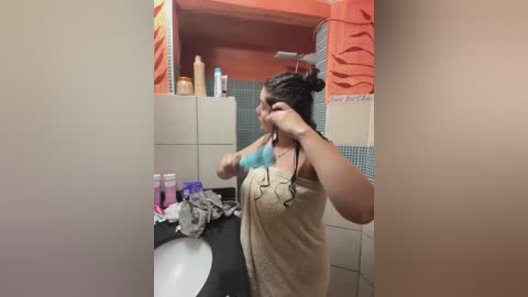 Media: A candid video of a woman with medium skin tone, wearing a beige towel, brushing her hair in a bathroom with orange and blue tiled walls, cluttered countertop, and toiletries.