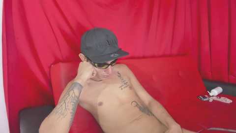 Media: Video of a shirtless, tattooed young man with a black cap and sunglasses reclining on a red couch against red curtains.