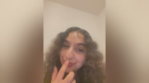 Media: A video of a young woman with curly, voluminous brown hair, light brown skin, and a playful expression, using her index finger to cover her lips. She has a slight smile and is wearing a black top.