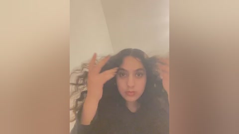 Media: Video of a young woman with long, dark hair, light brown skin, and a slender physique, wearing a black top, posing with both hands in her hair. Background features a beige wall and a patterned pillow.