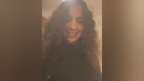 Media: Video of a smiling, curly-haired young woman with light brown skin, wearing a black top, standing in a dimly lit room with a blurred background.