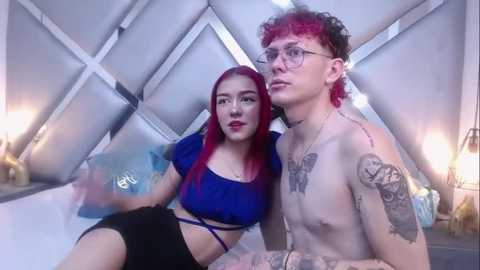 Media: Video of a young woman with red hair, wearing a blue crop top, and a tattooed man with curly red hair and glasses, in a modern, geometric room with white walls and lamps.