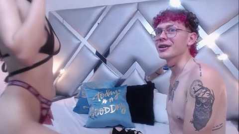 Media: Video of a young, light-skinned man with curly red hair, wearing glasses, and a tattoo on his right arm, standing in a modern, white-tiled bedroom with a patterned headboard.