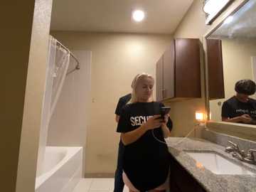 Media: Video of a blonde woman in a black \"SERVE\" t-shirt, focused on her smartphone, in a beige-tiled bathroom with a white bathtub, dark wooden cabinets, and a granite countertop.