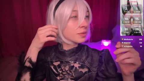 Video of a pale-skinned person in a white wig with black hairband, wearing a black lace top, holding a smartphone, set against a red background with a live stream screen.