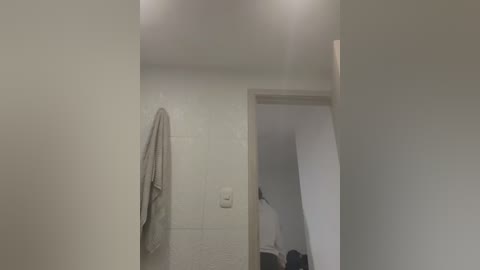 Video of a minimalist bathroom with off-white tiles, a beige towel hanging on a hook, and a narrow staircase leading to an upper floor.