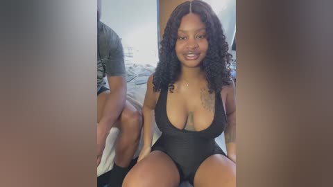 Video of a smiling, dark-skinned woman with curly hair, wearing a black, low-cut one-piece swimsuit, sitting on a bed with a man partially visible behind her.