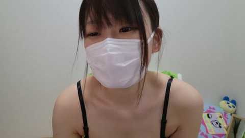 Media: Video of an East Asian woman with dark hair in a bun, wearing a white surgical mask and black spaghetti straps, standing against a plain white wall with a stuffed toy and a colorful toy box in the background.