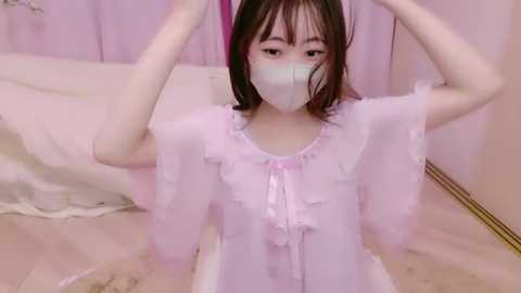 Media: Video of an Asian woman with straight black hair, wearing a pastel pink, frilly, see-through dress with a lace bodice and a face mask, sitting on a white bed, indoors.