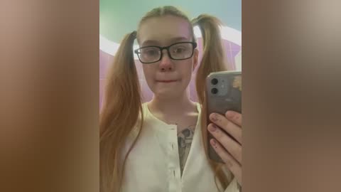 Media: A video of a young woman with long, light brown hair styled in pigtails, wearing black-framed glasses, a white shirt, and a tattoo on her chest. She's taking a selfie in a bathroom with tiled walls.