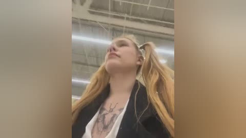 A video of a young woman with long, blonde hair in pigtails, wearing a black jacket over a white shirt with a deer tattoo, standing in an industrial setting with high ceilings and exposed beams.