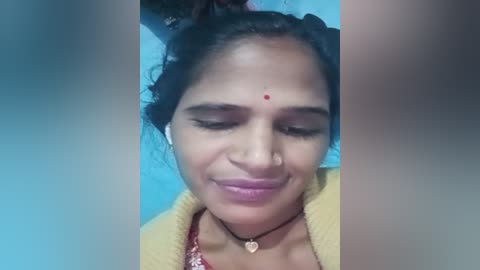 Media: Video of a South Asian woman with medium brown skin, wearing a yellow cardigan and a red bindi on her forehead. She has curly hair and a subtle smile.