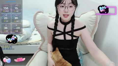 Video of a young Asian woman with black hair, glasses, and a black strappy top, sitting on a beige chair. She holds an orange cat. Background shows a white wall and a digital health tracker.