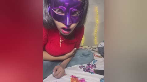 Media: A video of a woman with light skin, medium build, and medium breasts, wearing a purple mask, red top, and blue jeans, sitting on a bed with a colorful blanket, in a dimly lit room.