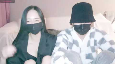 Media: Video of two Asian women, one in a black mask and black outfit, the other in a black mask, black hat, and white checkered pajamas, sitting closely on a bed with white sheets.