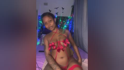 Media: Video of a tattooed, dark-skinned woman with medium-sized breasts, wearing a red lingerie set with bows, sitting on a bed with colorful string lights and bird decorations in the background.