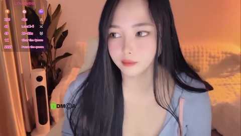 Media: A video of a young Asian woman with long black hair, light skin, and a slim physique, wearing a light blue blouse, seated indoors with a blurred background.