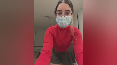 Media: A video of a young woman with light skin and long brown hair, wearing a red long-sleeve shirt, black skirt, and a blue face mask, standing in a modern, bright room with white walls and a red background.