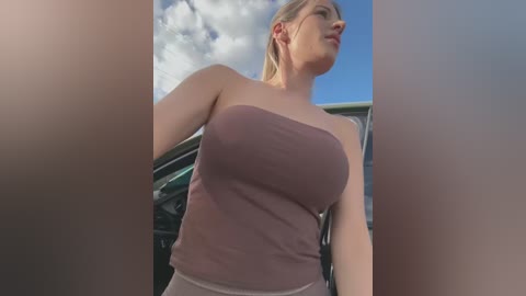 Media: Video of a curvy, light-skinned woman with blonde hair, wearing a strapless, maroon tank top, standing outdoors by a car with a blue sky background.