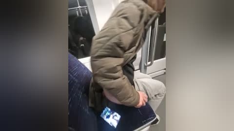 Media: Video of a person in a green jacket, light gray pants, and dark jeans, sitting on a train seat, reading a smartphone.