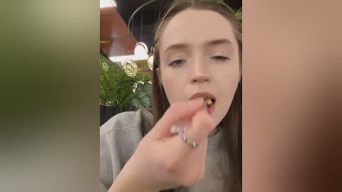 Media: Video of a young woman with straight, light brown hair and fair skin, wearing a gray sweater, licking a lollipop, surrounded by lush green plants and wooden furniture in a cozy indoor setting.