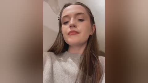 Media: Video of a young Caucasian woman with fair skin and long brown hair, wearing a light gray sweater, standing against a blurred beige background.