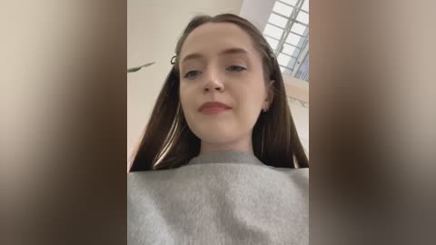 Media: A video of a young Caucasian woman with fair skin, straight brown hair, and a light gray sweater. She has a neutral expression, with a small, delicate nose and slightly parted lips. The background features a window with frosted glass and a hint of a plant.