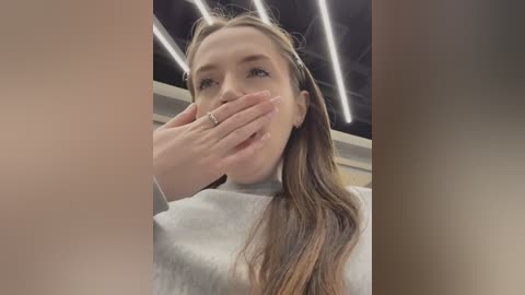 Media: A video of a young Caucasian woman with long brown hair, wearing a white sweater, covering her mouth in an indoor setting with modern, fluorescent lighting.