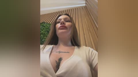 A video of a smiling, light-skinned woman with long brown hair and a tattoo of \"forever\" on her chest, wearing a beige ribbed top, standing in front of a wooden-paneled wall and green foliage.