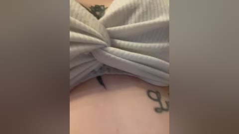 A close-up video of a person's stomach with a light grey ribbed top tied in a knot, revealing a black tattoo of a cursive word. The skin is pale, with a tattoo on the right side.